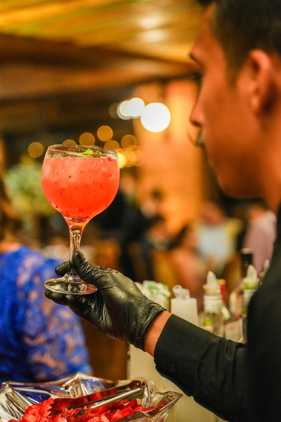 Bartender servindo um coquetel vermelho em taça alta, sob luz quente