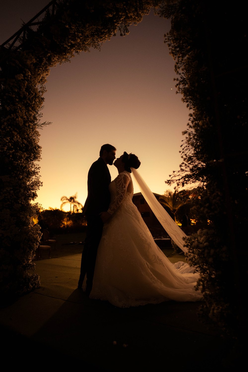 Casal em silhueta sob um arco de folhagem, contra o pôr do sol